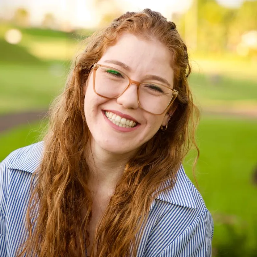 Retrato da neuropsicóloga Bruna Leticia Kunrath sorrindo ao ar livre, em enquadramento quadrado, com camisa listrada azul e branca e óculos.